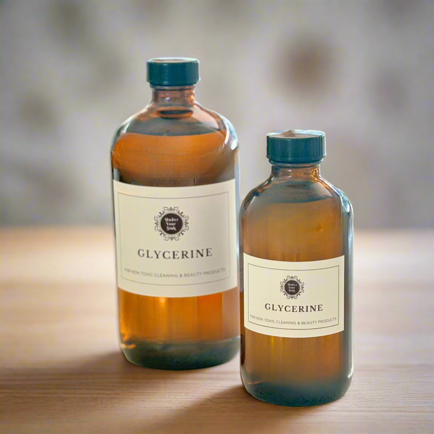 A collection of 2 amber glass bottles in 500ml and 250ml sizes. Each has a simple white label with Under your Sink Logo and description Glycerine – For non toxic cleaning and beauty products. The bottles are set in a kitchen with a blurred background. It is a product image for website as this is for sale in various sizes, online with Australia wide delivery.