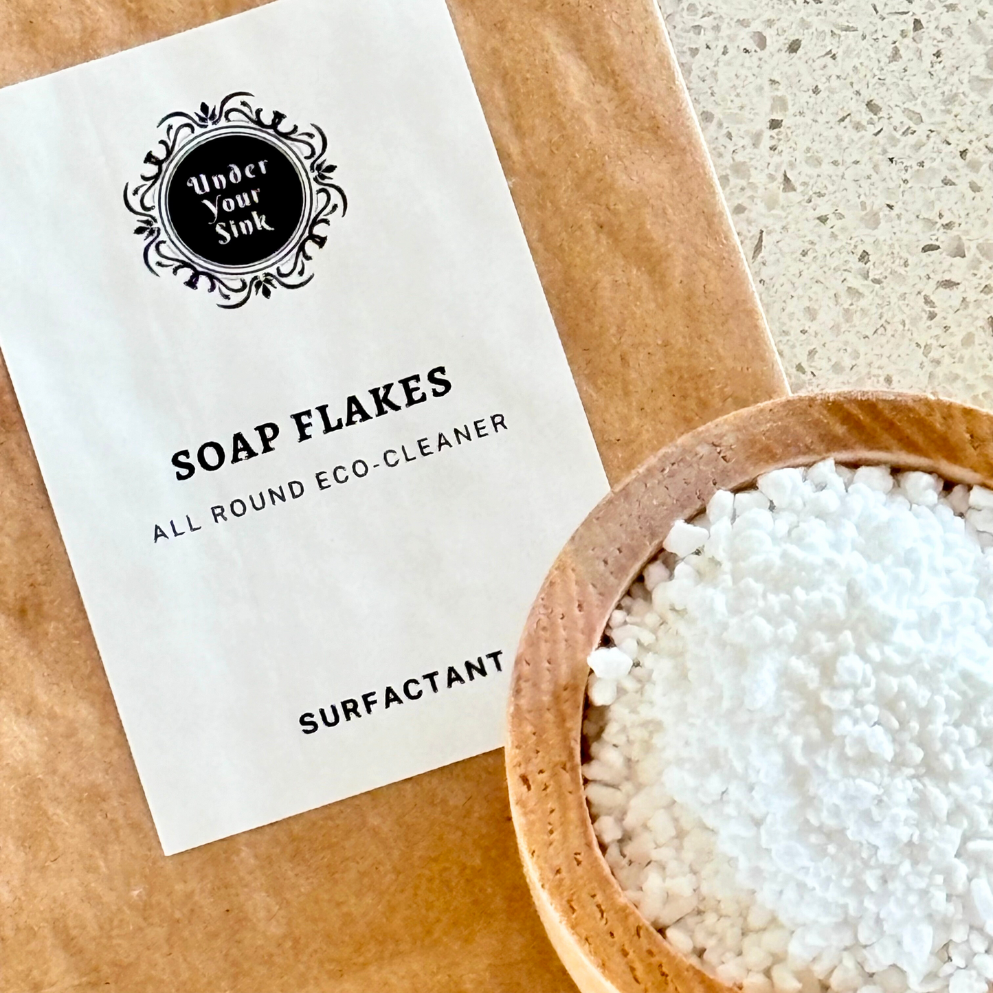 A kraft paper earth bag with simple white label with Under your Sink Logo and description Soap Flakes- all round Eco-cleaner – surfactant. There is a little wooden bowl with white ground up coconut soap flakes for DIY cleaning and beauty products. This is a product image for website as this is for sale in various sizes, online with Australia wide delivery.
