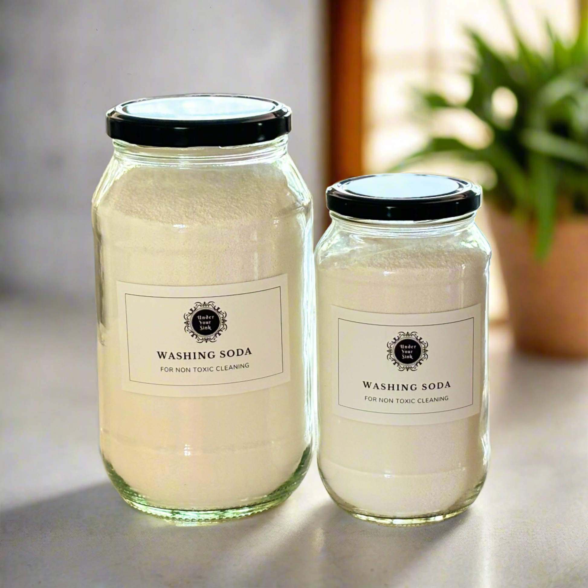 Two clear glass jars, one larger (2Litres) and one smaller (1litre). They both have simple white label with Under your Sink Logo and description Washing Soda - all round Eco-cleaner – heavy duty alkali. They are filled with white crystalline washing soda granules inside. Background is of a kitchen and its a clean image to show green cleaning. This is a product image for website as this is for sale in various sizes, online with Australia wide delivery.