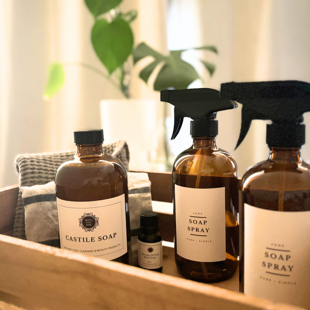All purpose spray kit with amber glass spray bottles and amber glass bottle of Castile soap in a wooden box with some cleaning rags. 