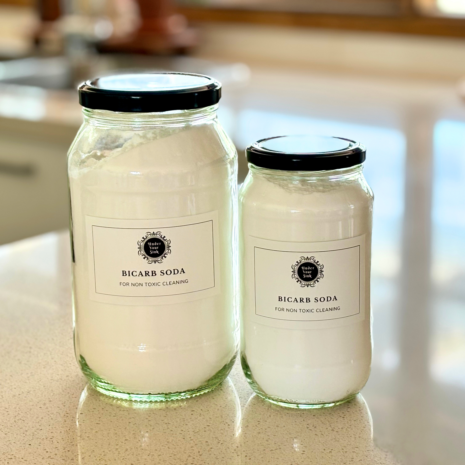 Two clear glass jars, one larger (2Litres) and one smaller (1litre). They both have simple white label with Under your Sink Logo and description Bicarb- all round Eco-cleaner – mild strength alkali. They are filled with white crystalline Bicarb granules inside. Background is of a kitchen and its a clean image to show green cleaning. This is a product image for website as this is for sale in various sizes, online with Australia wide delivery.