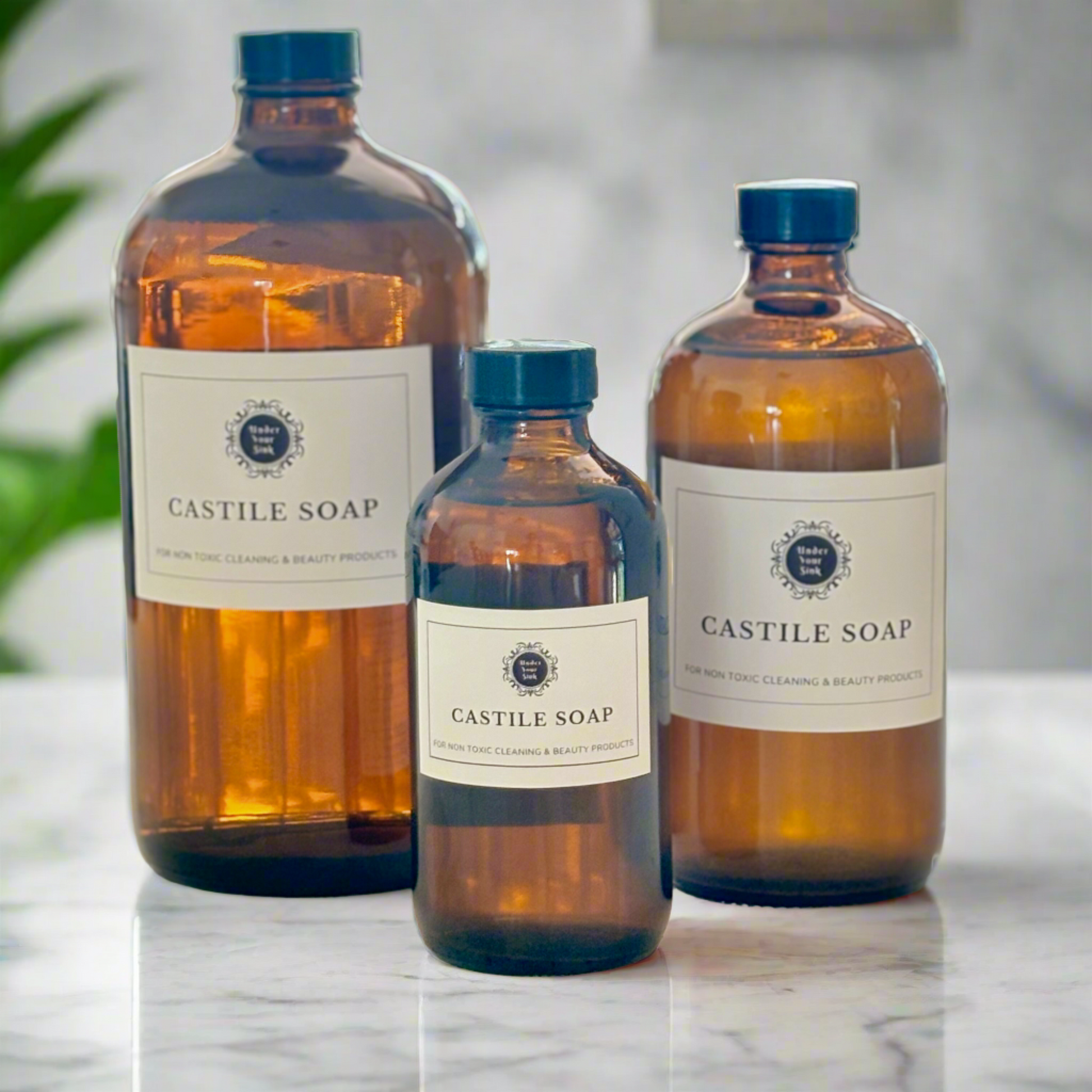 A collection of 3 amber glass bottles in 1litre, 500ml and 250ml sizes. Each has a simple white label with Under your Sink Logo and description Castile Soap – For non toxic cleaning and beauty products. The bottles are set in a kitchen with a blurred background for effect. It is a product image for website as this is for sale in various sizes, online with Australia wide delivery.