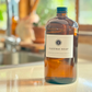 An amber glass bottle with a white label with Under your Sink Logo and description Castile Soap – For non toxic cleaning and beauty products. The bottles are set in a kitchen with a blurred background for effect. It is a product image for website as this is for sale in various sizes, online with Australia wide delivery.