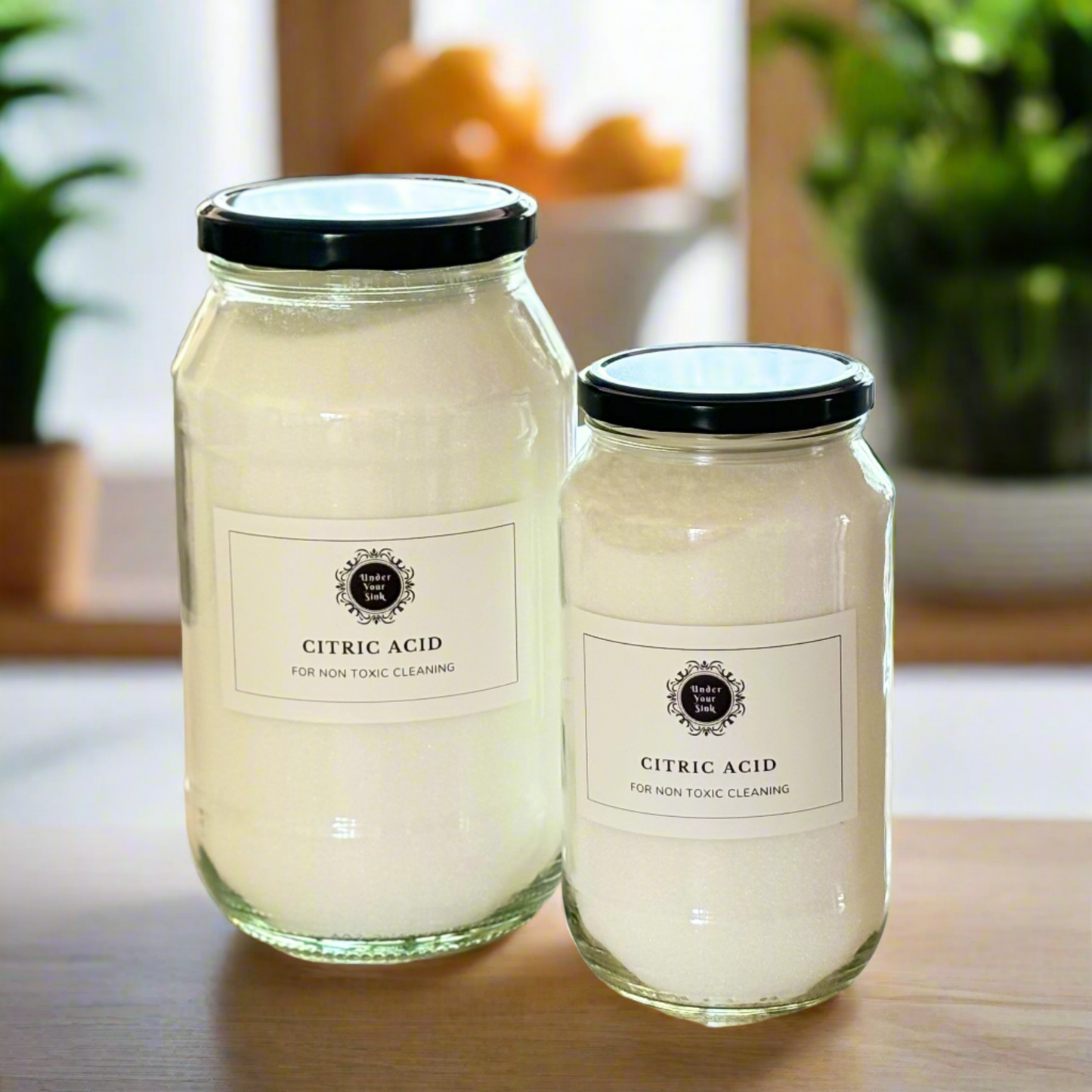 Two clear glass jars, one larger (2Litres) and one smaller (1litre). They both have simple white label with Under your Sink Logo and description Citric Acid- all round Eco-cleaner – acidic solvent. They are filled with whitish clear crystal like Citric Acid for natural cleaning.  Background is of a kitchen and its a clean image to show green cleaning. This is a product image for website as this is for sale in various sizes, online with Australia wide delivery.