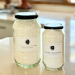 Two clear glass jars, one larger (2Litres) and one smaller (1litre). They both have simple white label with Under your Sink Logo and description Soap Flakes- all round Eco-cleaner – surfactant. They are filled with white ground up coconut soap flakes for DIY cleaning and beauty products. inside. Background is of a kitchen and its a clean image to show green cleaning. This is a product image for website as this is for sale in various sizes, online with Australia wide delivery.