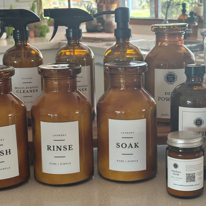 A group of amber glass laundry detergent bottles and spray bottles with white labels, arranged on a kitchen counter in front of a window.