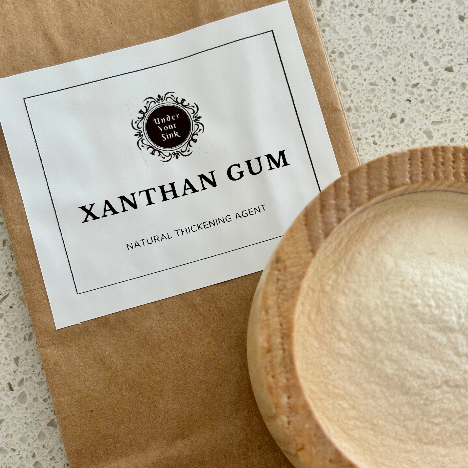 A kraft paper earth bag with a simple white label with Under your Sink Logo and description Xanthan Gum – natural thickening agent. There is also a small wooden bowl with the very fine xanthan gum powder that is almost beige in colour. It is a product image for website as this is for sale in various sizes, online with Australia wide delivery.