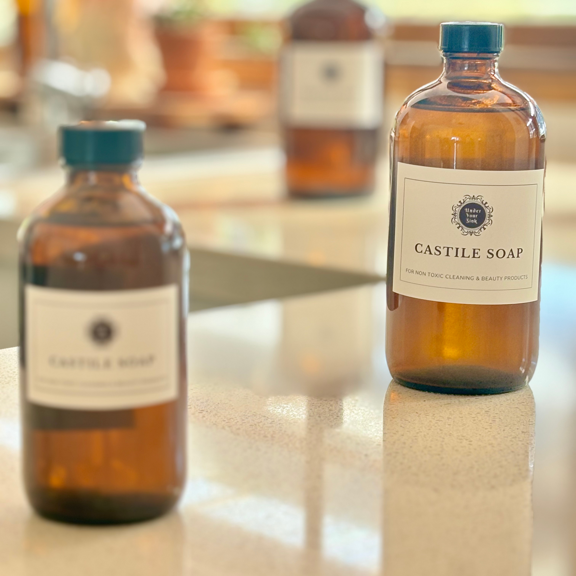 A collection of 3 amber glass bottles in 1litre, 500ml and 250ml sizes. Each has a simple white label with Under your Sink Logo and description Castile Soap – For non toxic cleaning and beauty products. The bottles are set in a kitchen with a blurred background a focus on the 500ml bottle. It is a product image for website as this is for sale in various sizes, online with Australia wide delivery.