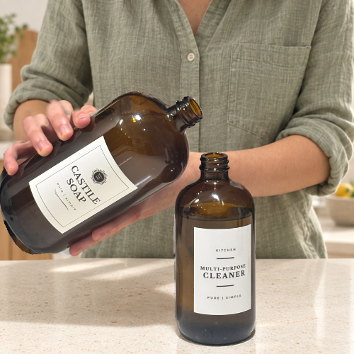 A person in a sage green shirt holds an amber glass bottle labeled 'CASTLE SOAP' over a countertop next to another amber glass bottle labeled 'MULTI-PURPOSE CLEANER' by Under Your Sink