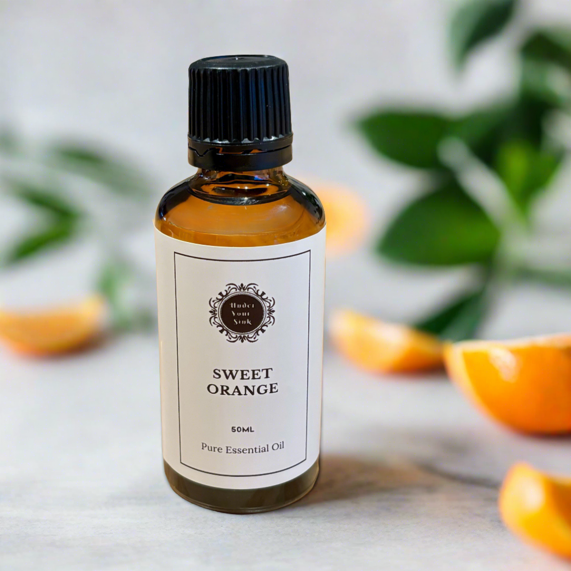Amber glass essential oil bottle with white label with Under Your Sink logo and description SWEET ORANGE pre essential oil. Has blurred images of oranges in background