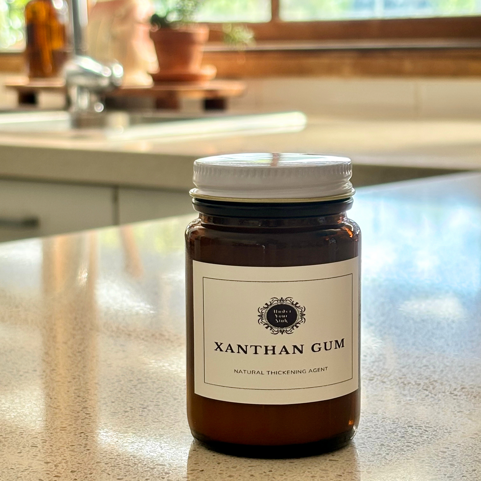 An amber glass jar with a simple white label with Under your Sink Logo and description Xanthan Gum – natural thickening agent. Set in a kitchen with blurred background and focus on the jar. It is a product image for website as this is for sale in various sizes, online with Australia wide delivery.