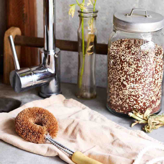 An EcoMax dish brush with a wooden handle and brown bristles resting on a beige cloth, alongside a glass jar filled with grains and a kitchen faucet in the background.